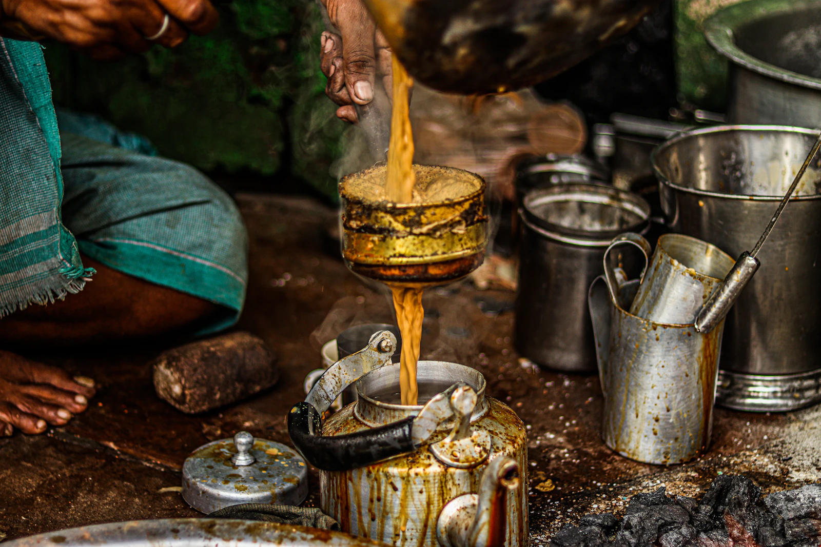 A chai vendor in Varanasi straining tea from a pan into a kettle over a coal fire, surrounded by steel pots and cups
